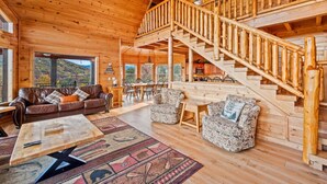 A spacious wooden cabin with a central living area, a dining table, and a staircase leading to an upper level.