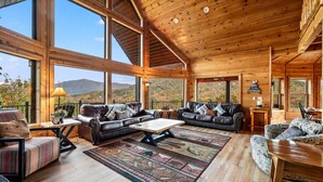 A spacious cabin with large windows offering mountain views, a cozy living area with leather sofas and a coffee table, and wooden flooring.