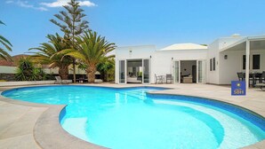 A swimming pool with a curved edge, surrounded by a patio area with chairs and tables. There are palm trees and a building with glass doors and a white exterior.