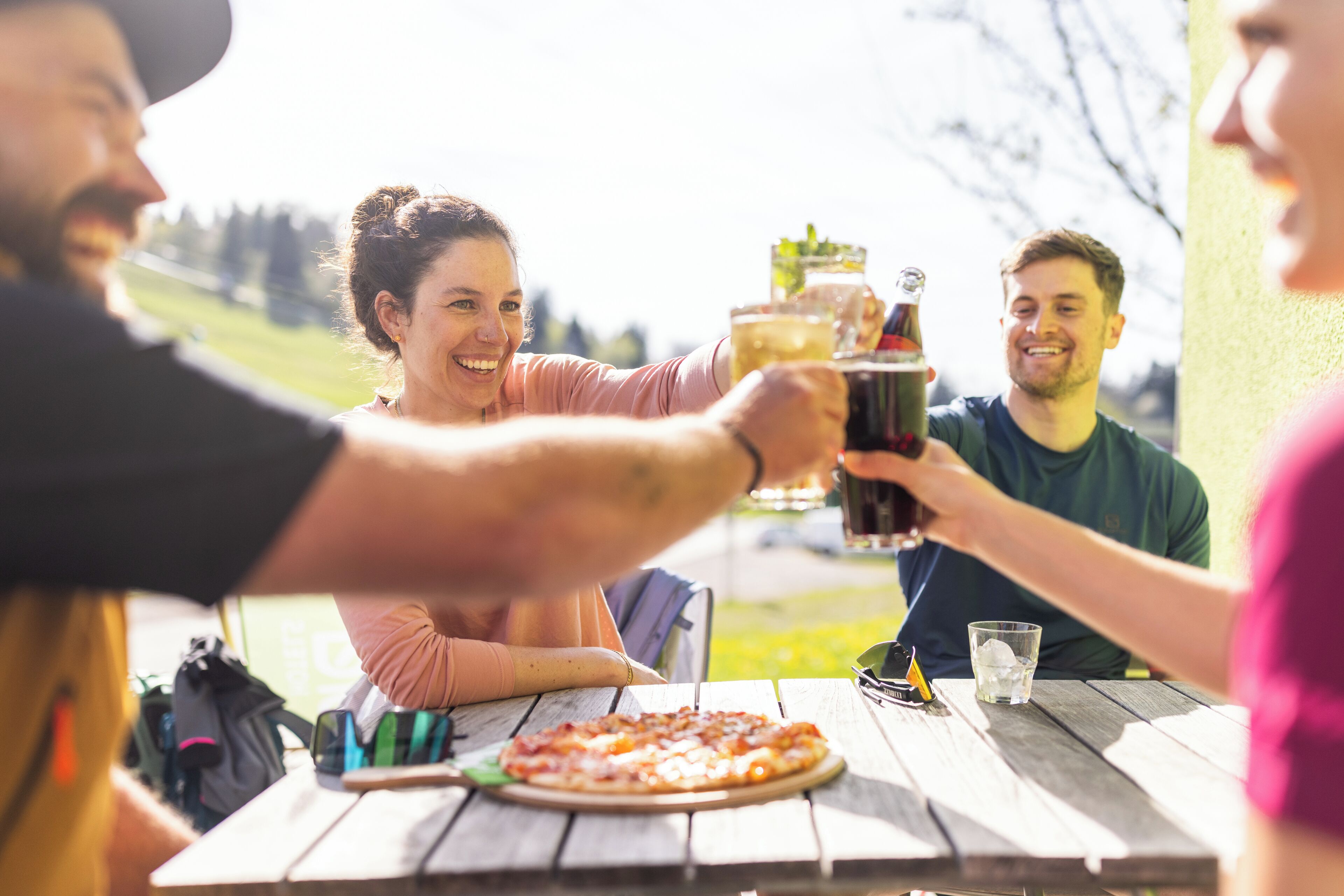 Un groupe de personnes trinquant autour d’une table en bois, devant une pizza et des boissons.