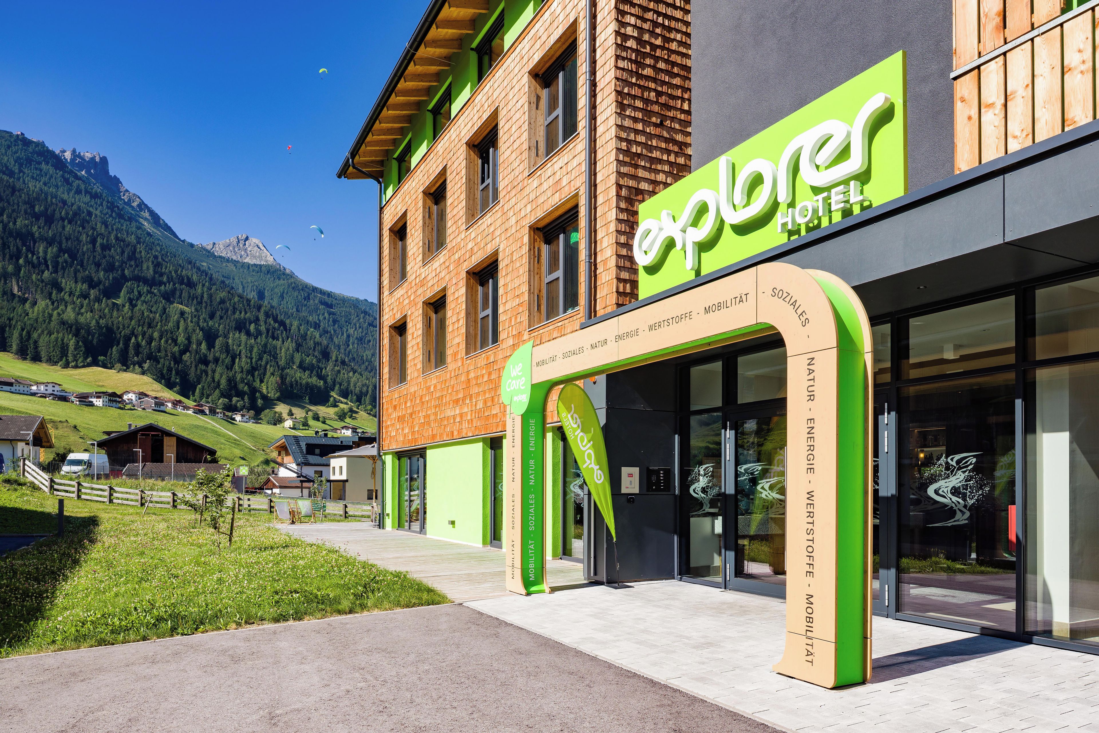 Un hotel con fachada verde y marrón, cielo despejado y montañas al fondo.