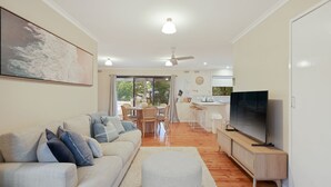 Een woonkamer met een bank, poef en een televisie op een houten dressoir. Er is een zithoek met een tafel en stoelen, en een keuken met een koelkast en een televisie.