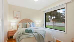 Een slaapkamer met een groot raam, een bed met opgerolde handdoeken, een nachtkastje met een lamp, en een lijst met een afbeelding aan de muur.