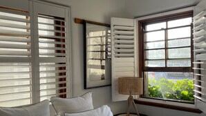 Un dormitorio con cama, almohadas blancas, una lámpara de noche, una ventana con persianas y vistas al exterior con vegetación.