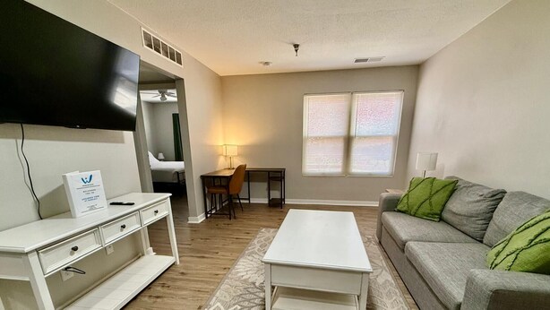 A modern living room with a flat-screen TV, a grey sofa with green cushions, a white coffee table, and a desk with a chair.