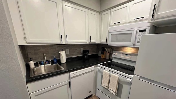 A compact kitchen with white cabinets, a black countertop, a dishwasher, an oven, and a microwave. - Modern Apartment Near Aksarben Village & Dining (Omaha)
