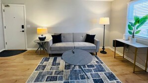 Un salon avec un canapé gris, une table basse ronde, un tapis à motifs, une lampe sur pied et une plante en pot près d’une fenêtre.