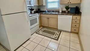 Une cuisine avec des meubles de couleur beige, un réfrigérateur blanc, une cuisinière, un lave-vaisselle et une fenêtre avec des stores.