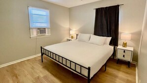 Une chambre à coucher avec un lit, deux tables de chevet, un ventilateur de plafond, une fenêtre avec des stores et un rideau foncé.
