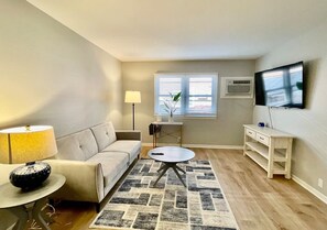 Una sala de estar con sofá, una mesita redonda, un televisor montado en la pared y una pequeña mesa auxiliar con lámpara.