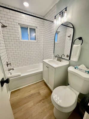 Un baño con paredes de azulejos blancos, una bañera, un lavamanos, un inodoro y un espejo.