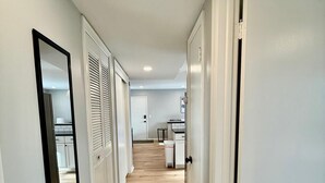 A hallway with white walls and wooden flooring, leading to a kitchen area with cabinets and a countertop.