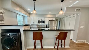 A modern kitchen with a central island, two bar stools, a washing machine, and pendant lighting.