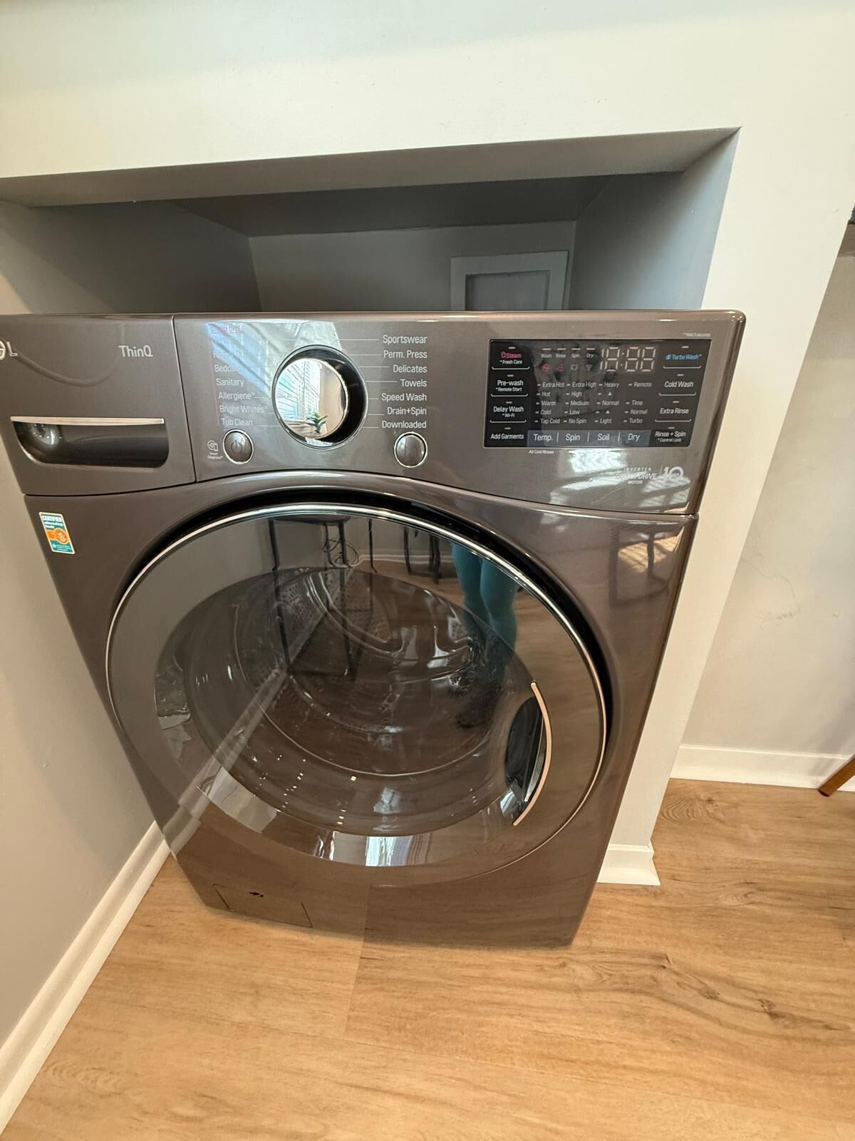 A front-loading washing machine with a control panel, located in a room with wooden flooring and white walls.