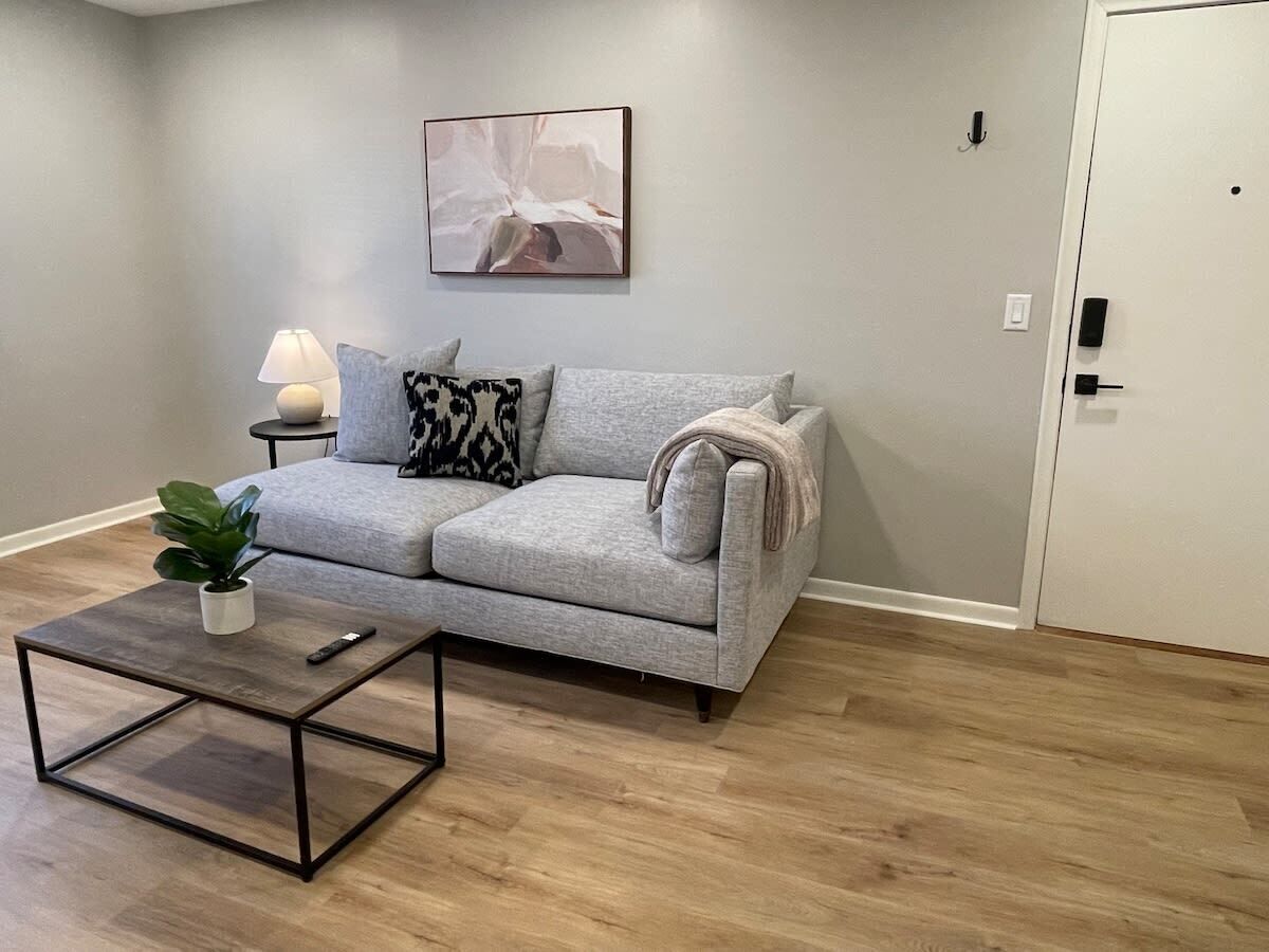A living room with a grey sofa, a small side table with a lamp, a coffee table, and a plant.