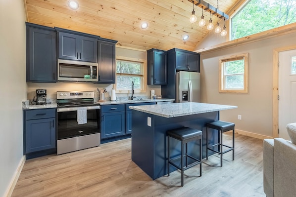 A modern kitchen with dark cabinets, stainless steel appliances, and a central island with bar stools. - The Falcons Nest- Hot Tub - Fire Pit - Large Covered Porch (Bryson City)