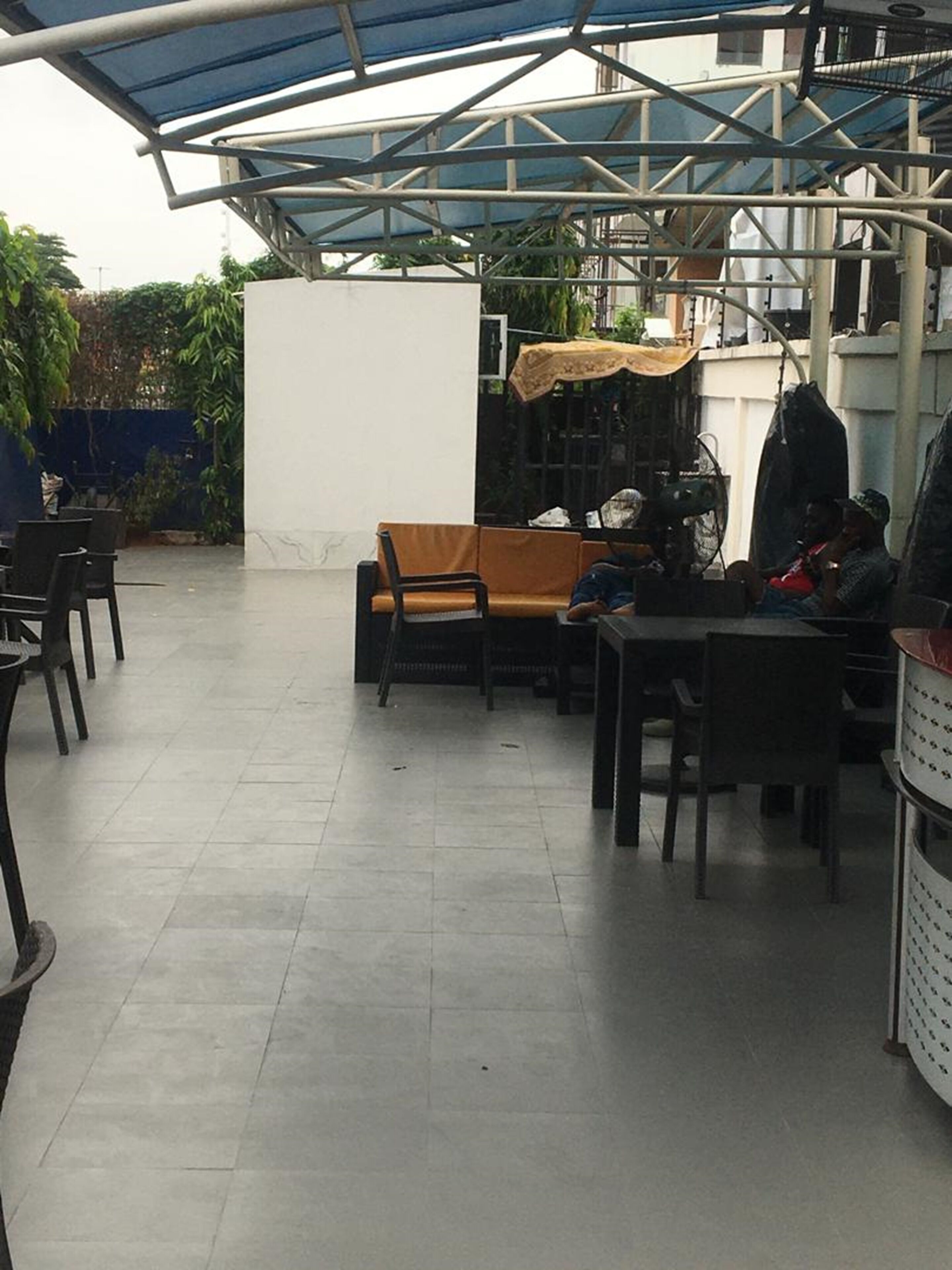 An outdoor seating area with tables and chairs, a covered section with a blue roof, and a white wall in the background.