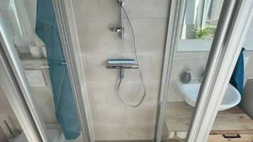 A glass-enclosed shower stall with a handheld showerhead, a wall-mounted faucet, and a small shelf.