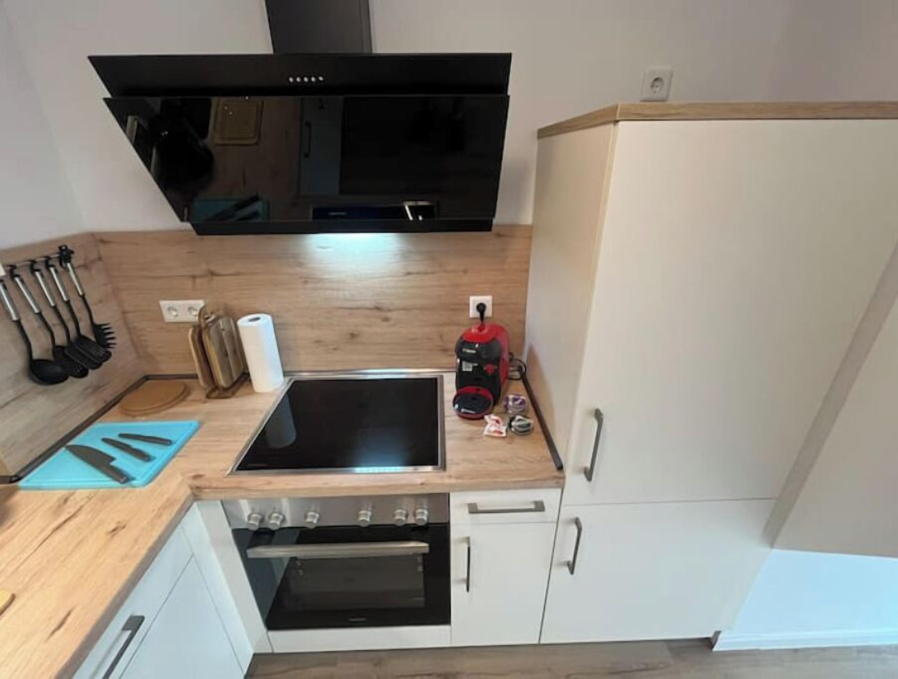 A compact kitchen with a built-in oven, a stovetop, and a microwave. There are utensils hanging on the wall, a roll of paper towels, and a vacuum cleaner on the counter.