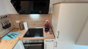 A compact kitchen with a built-in oven, a stovetop, and a microwave. There are utensils hanging on the wall, a roll of paper towels, and a vacuum cleaner on the counter.