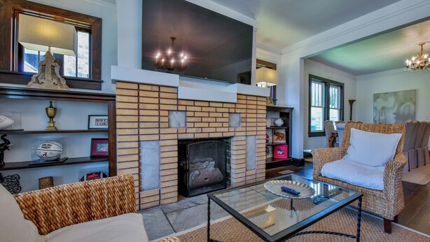 A living room with a brick fireplace, a flat-screen TV above it, a glass coffee table, wicker furniture, and a bookshelf with various items. - Heisman House - Book for football season!  Walk to Stadium! (Tuscaloosa)