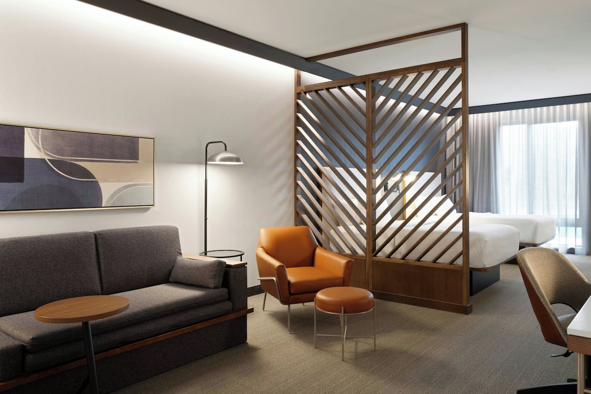 A modern hotel room with a grey sofa, an orange armchair, a small round table, and a bed with white linens.