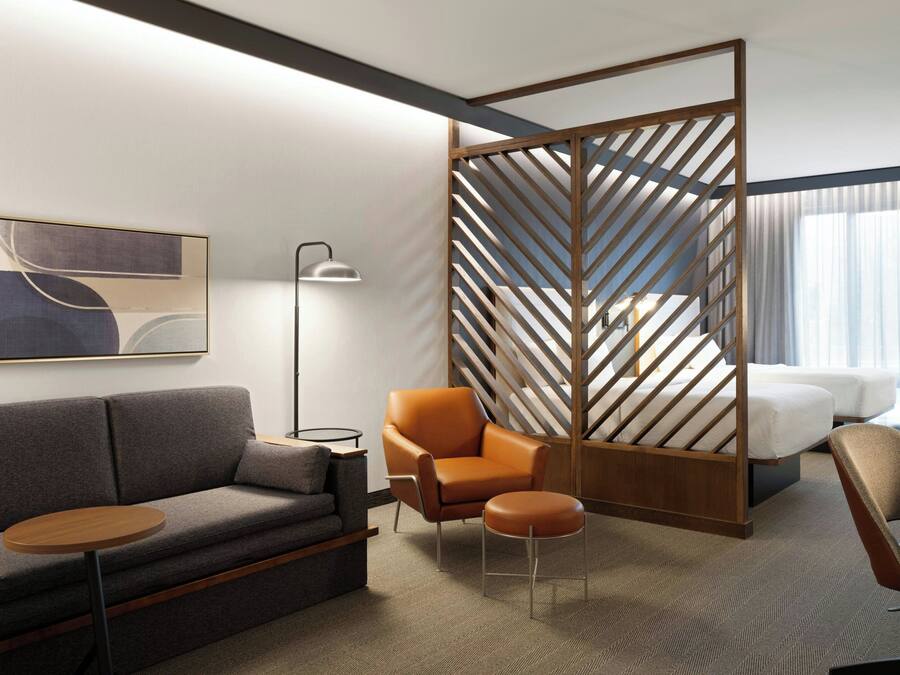 A modern hotel room with a grey sofa, an orange armchair, a small round table, and a bed with white linens.