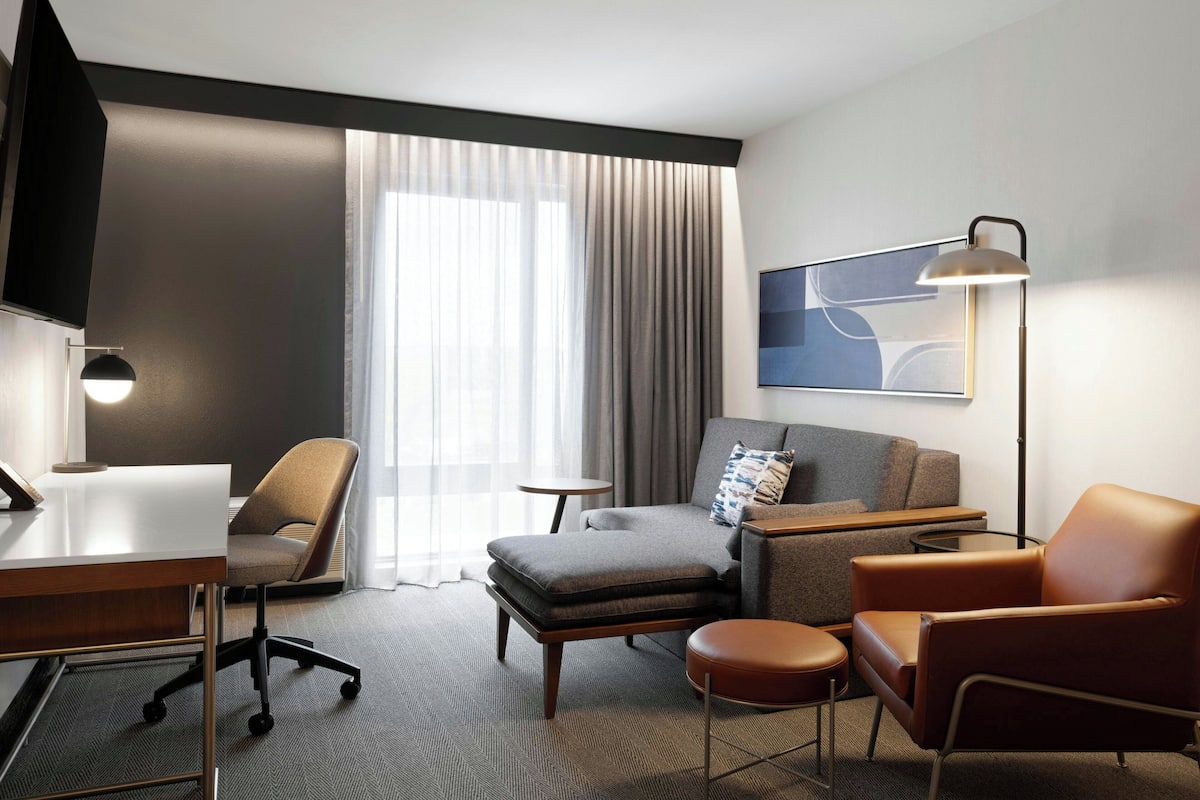 A modern hotel room with a desk, two armchairs, a sofa, a TV, a lamp, and a large window with curtains.