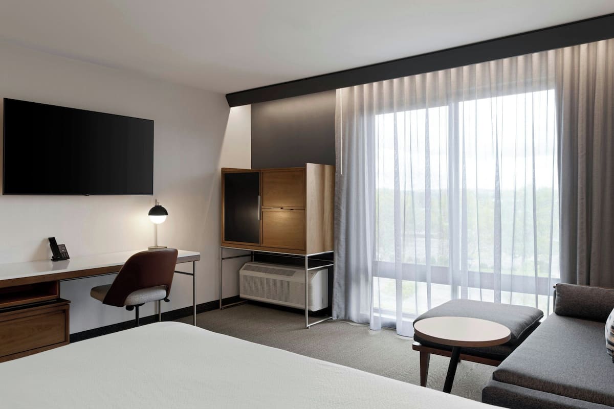A hotel room with a bed, a desk with a chair, a TV, a heater, a sofa, and a large window with curtains.