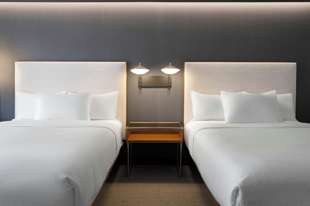A hotel room with two beds, a nightstand, and wall-mounted lighting.