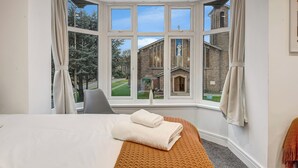 A hotel room with a large bay window, a bed with a white duvet and a brown throw, a chair, and a view of a building with a statue. - Large 4BR Cowley House, 4 Bath, Sleeps 10 with Parking (Oxford)
