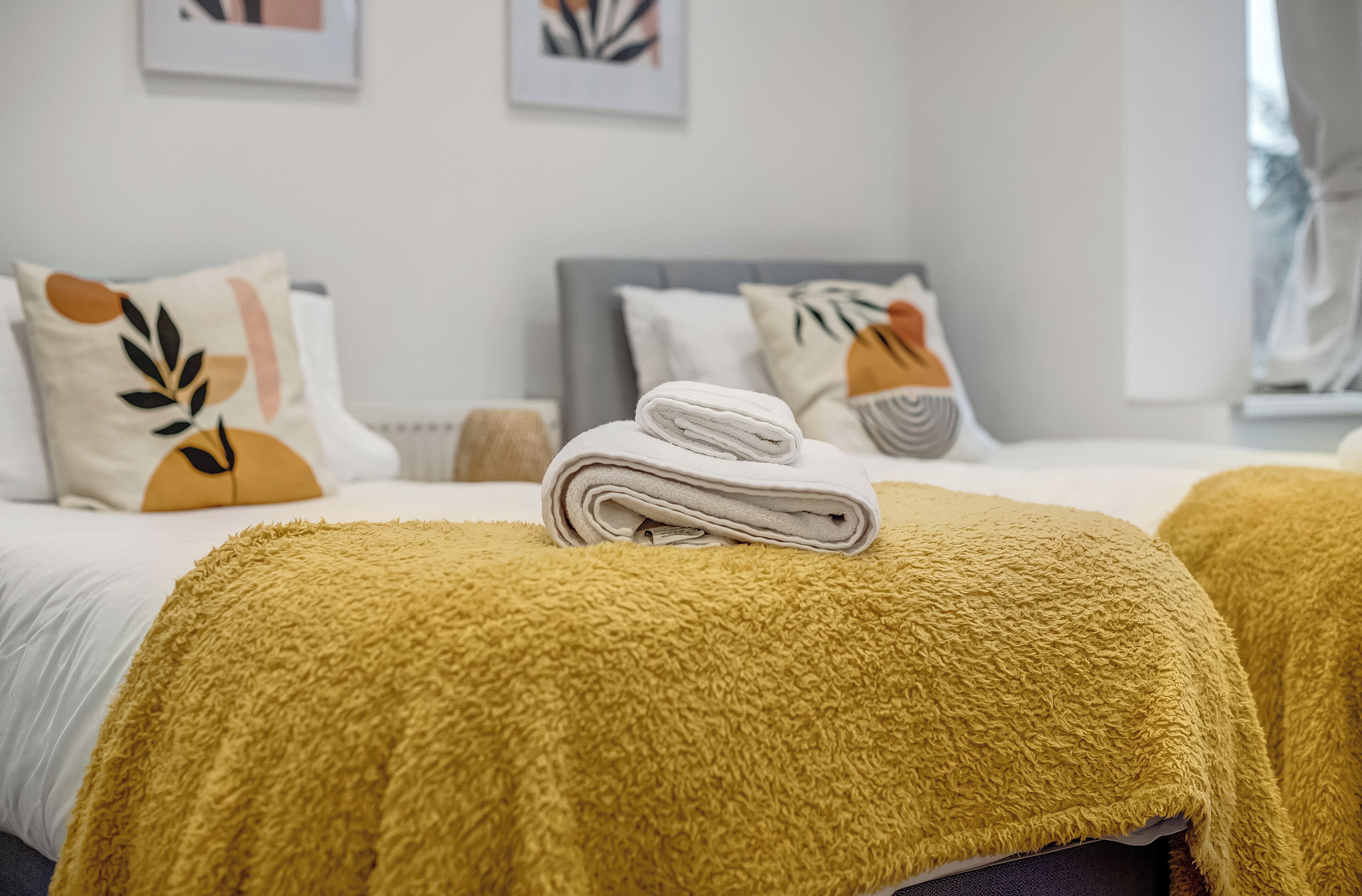 Una cama bien hecha con una toalla blanca doblada, un plaid color mostaza y almohadones con diseños de hojas.