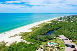 An aerial view of a coastal area with a beach, a swimming pool, and surrounding greenery.