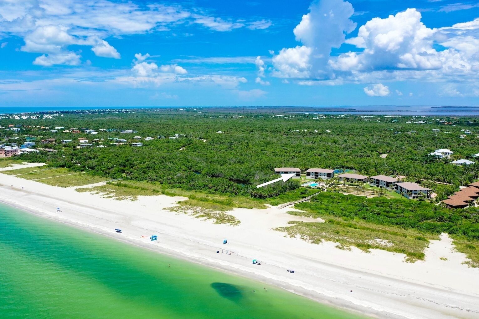An aerial view of a coastal area with a beach, a resort with multiple buildings, and surrounding greenery.