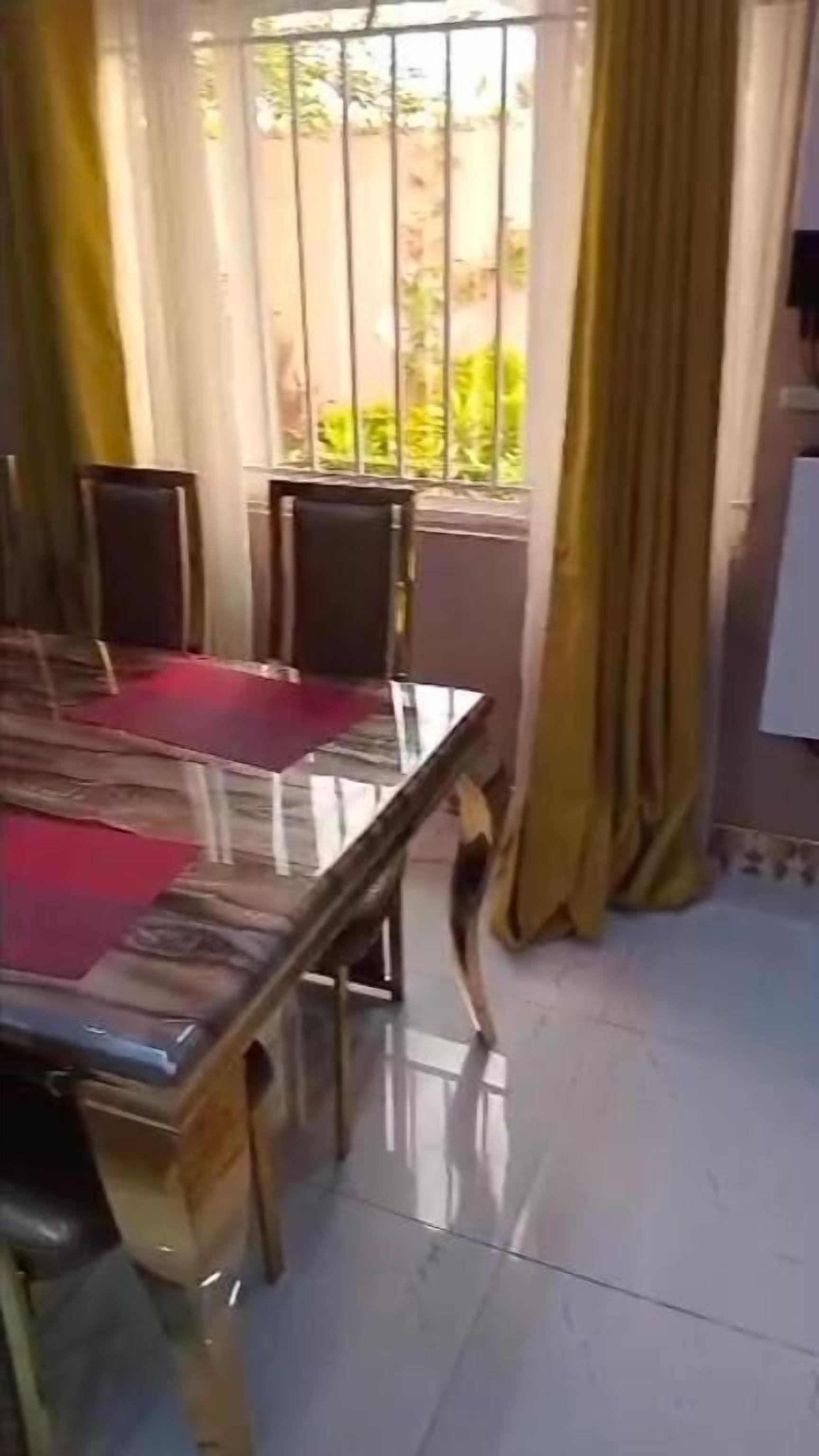 Un comedor con mesa y sillas de cristal, cortinas amarillas y vistas al exterior con vegetación.
