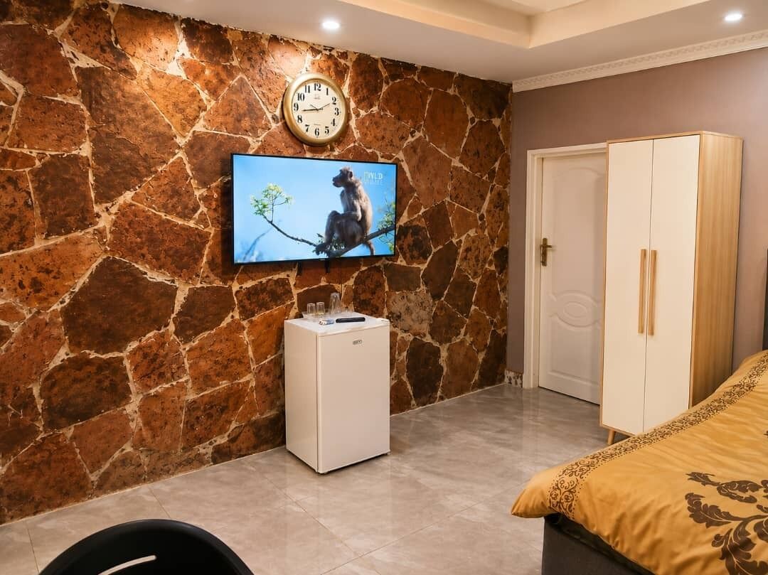 A room with a stone wall, a television mounted above it, a white mini-fridge, a wardrobe, and a bed with a patterned bedspread.