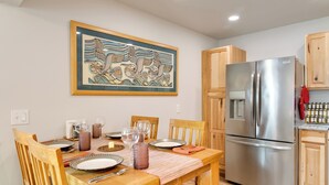 Une table à manger dressée avec des assiettes, des verres et des couverts, un réfrigérateur en acier inoxydable, des meubles de rangement en bois et une œuvre d’art accrochée au mur.