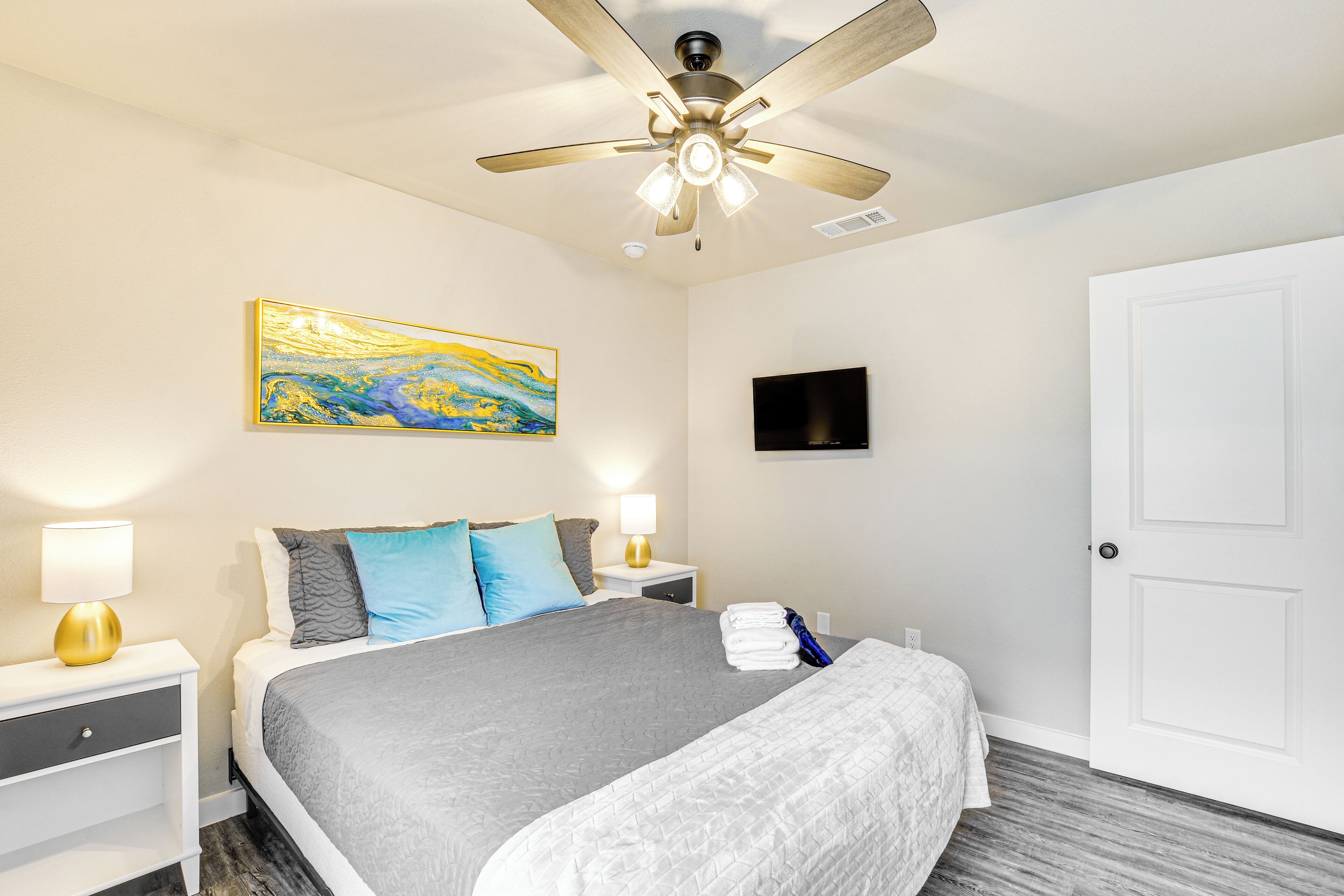 A neatly arranged bedroom with a bed, nightstands, a ceiling fan, a painting, a TV, and a door.