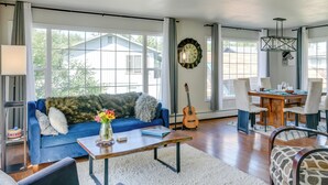 A spacious living room with a blue sofa, a coffee table, a dining area with chairs, a guitar, and a large window with curtains. - Explore Chugach & Beaches! Anchorage Adventure Pad (Anchorage)