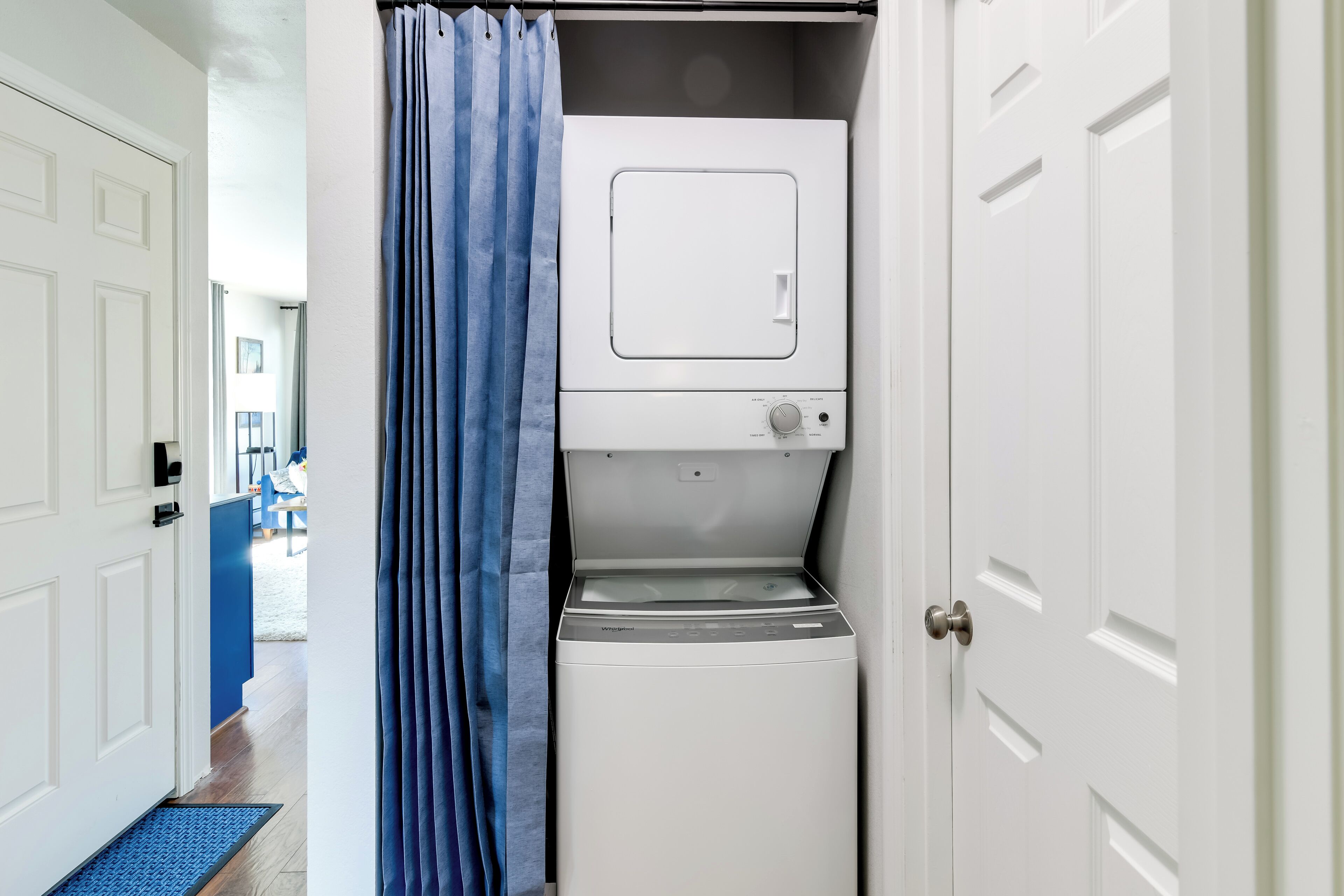 Une petite buanderie équipée d’un lave-linge et d’un sèche-linge, avec un rideau de douche bleu et une porte donnant sur un couloir.