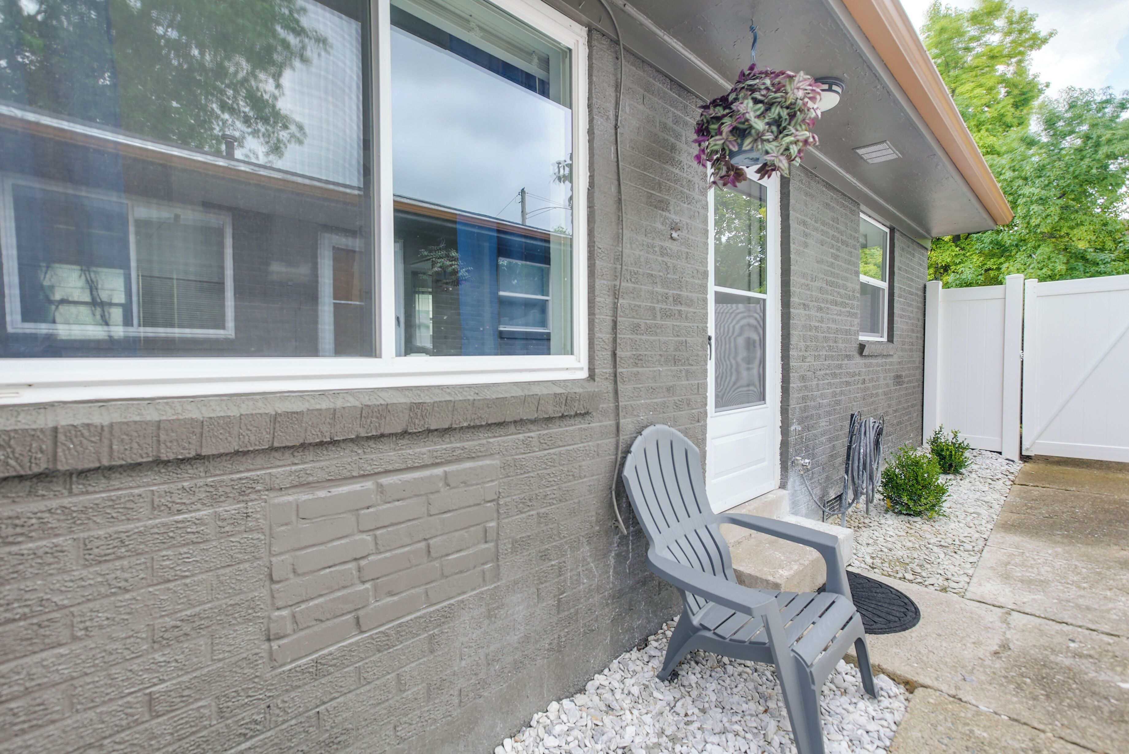 A house with a brick exterior, a window, a gray Adirondack chair, and a hanging flower basket.
