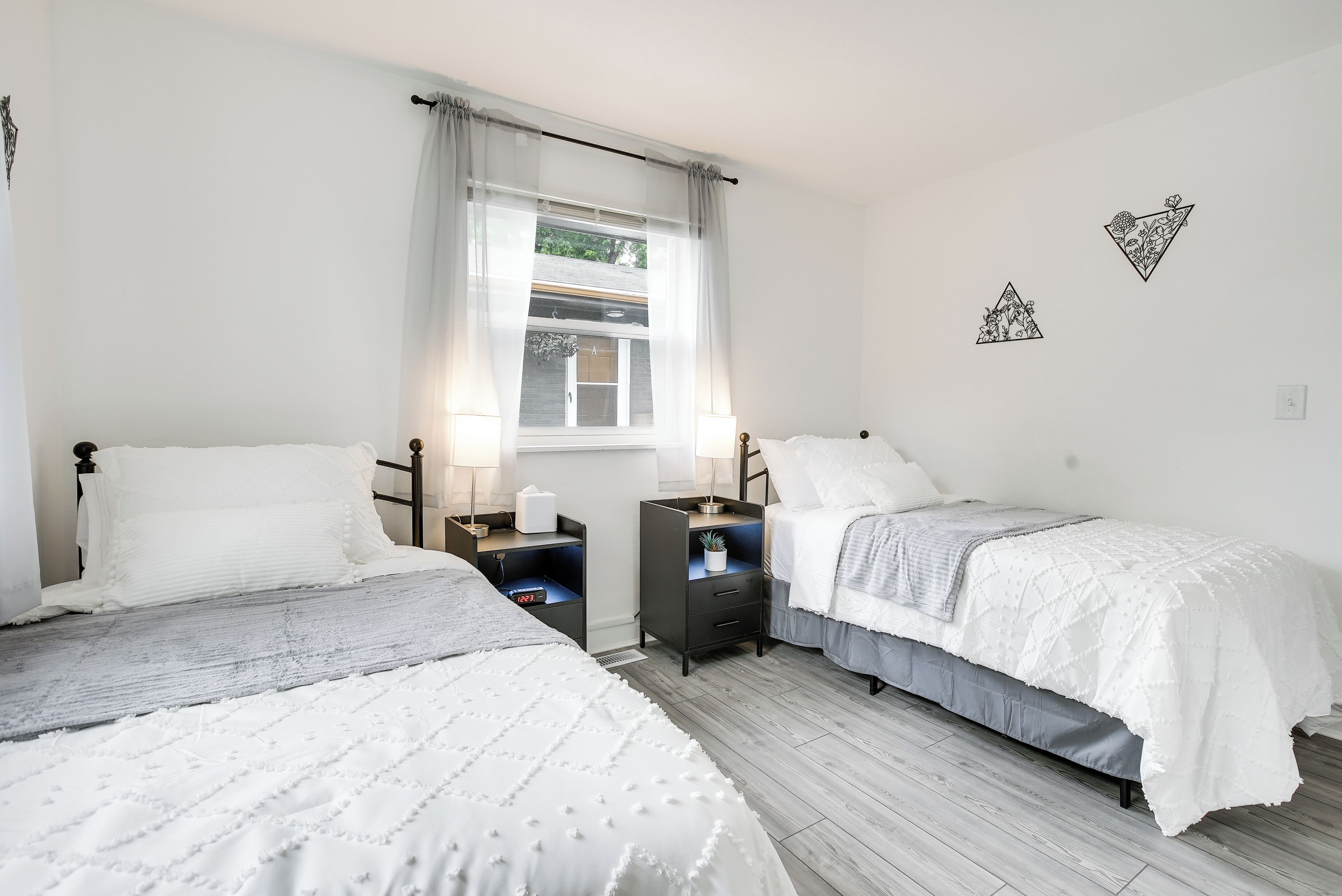 A bedroom with two beds, a window with sheer curtains, bedside tables with lamps, and wall art.