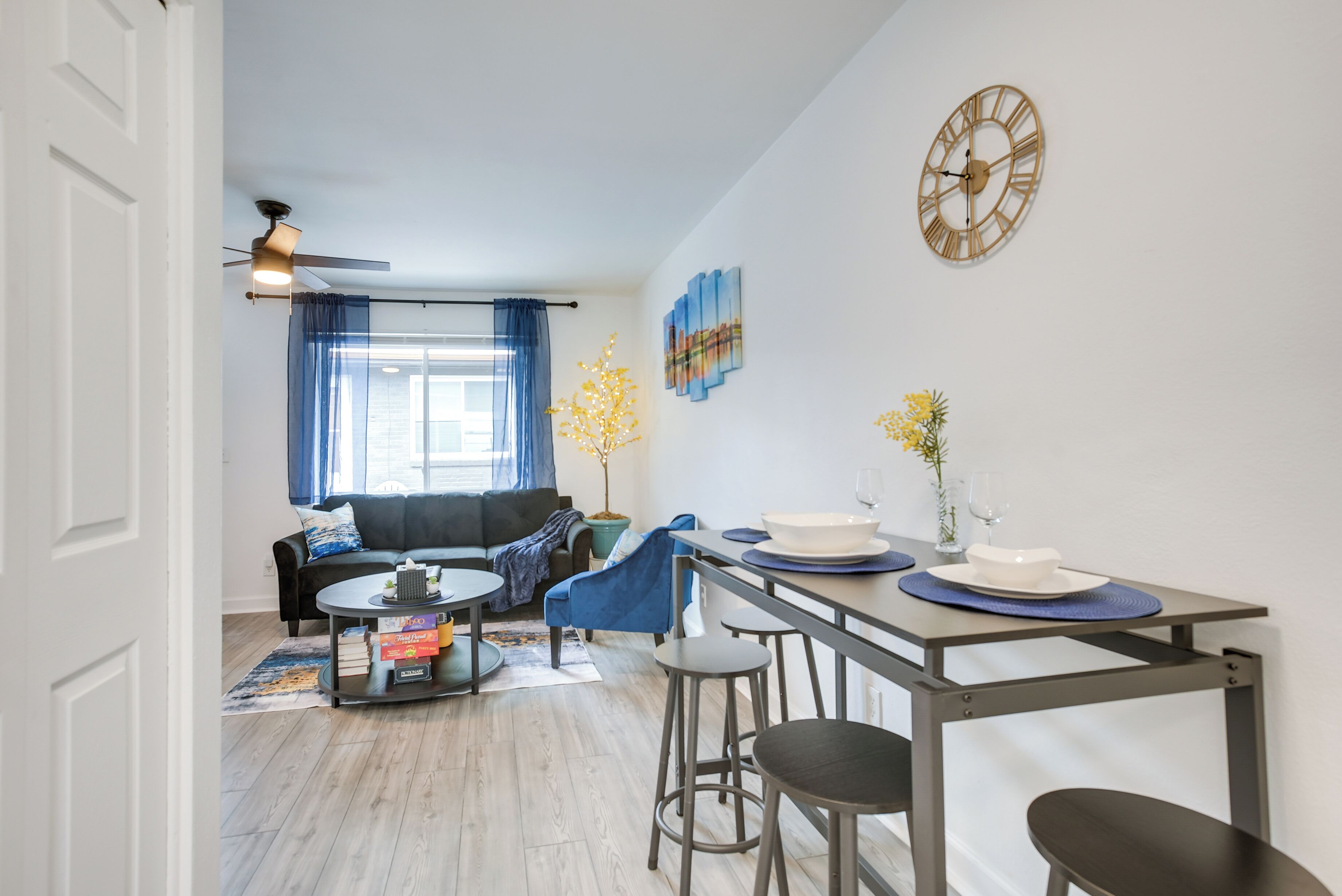 Ein modernes Wohnzimmer mit einer Couch, einem Esstisch mit Barhocker, einem Deckenventilator und einer Wanduhr.