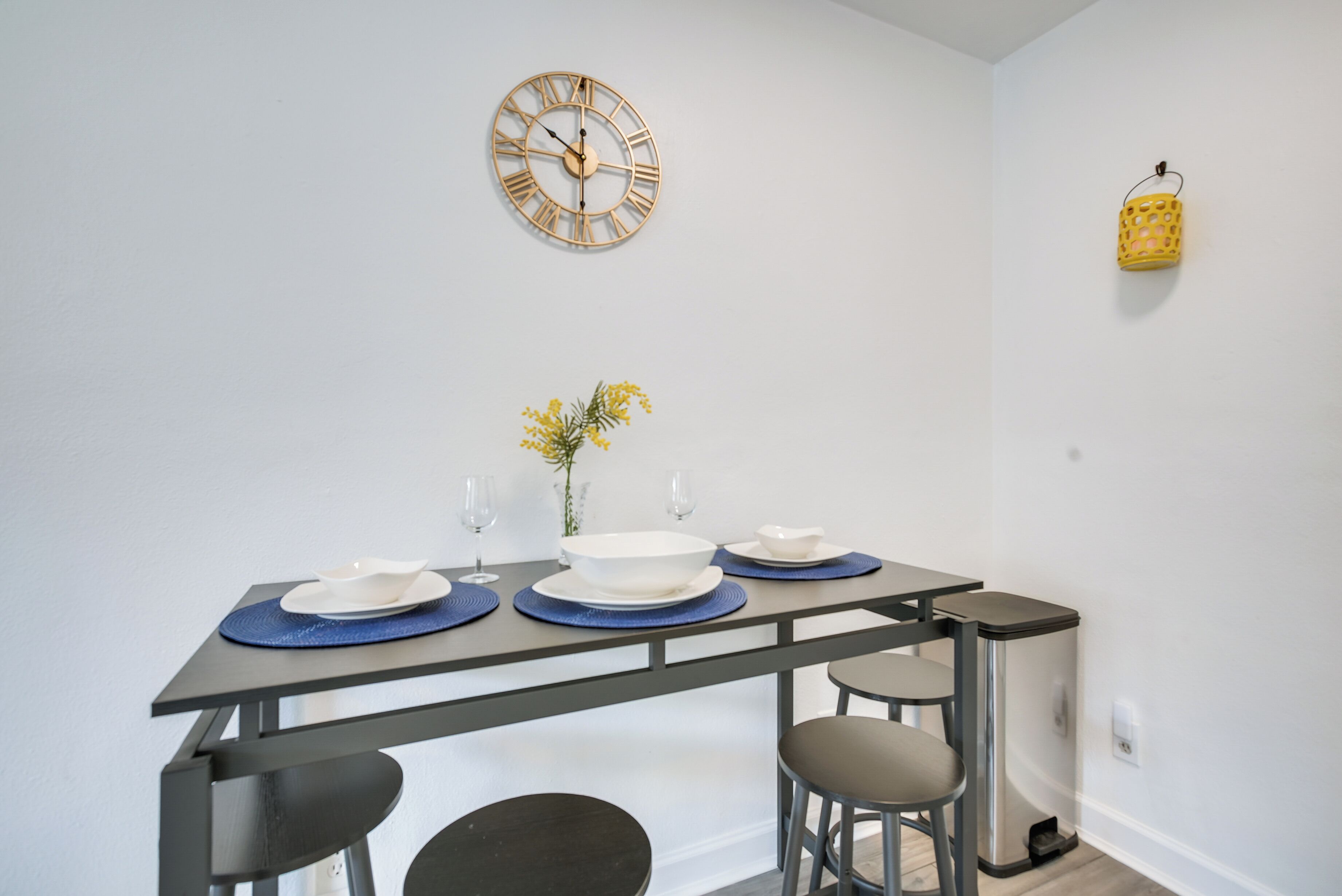Un comedor con una mesa preparada para cuatro personas, un reloj de pared y un pequeño adorno amarillo.