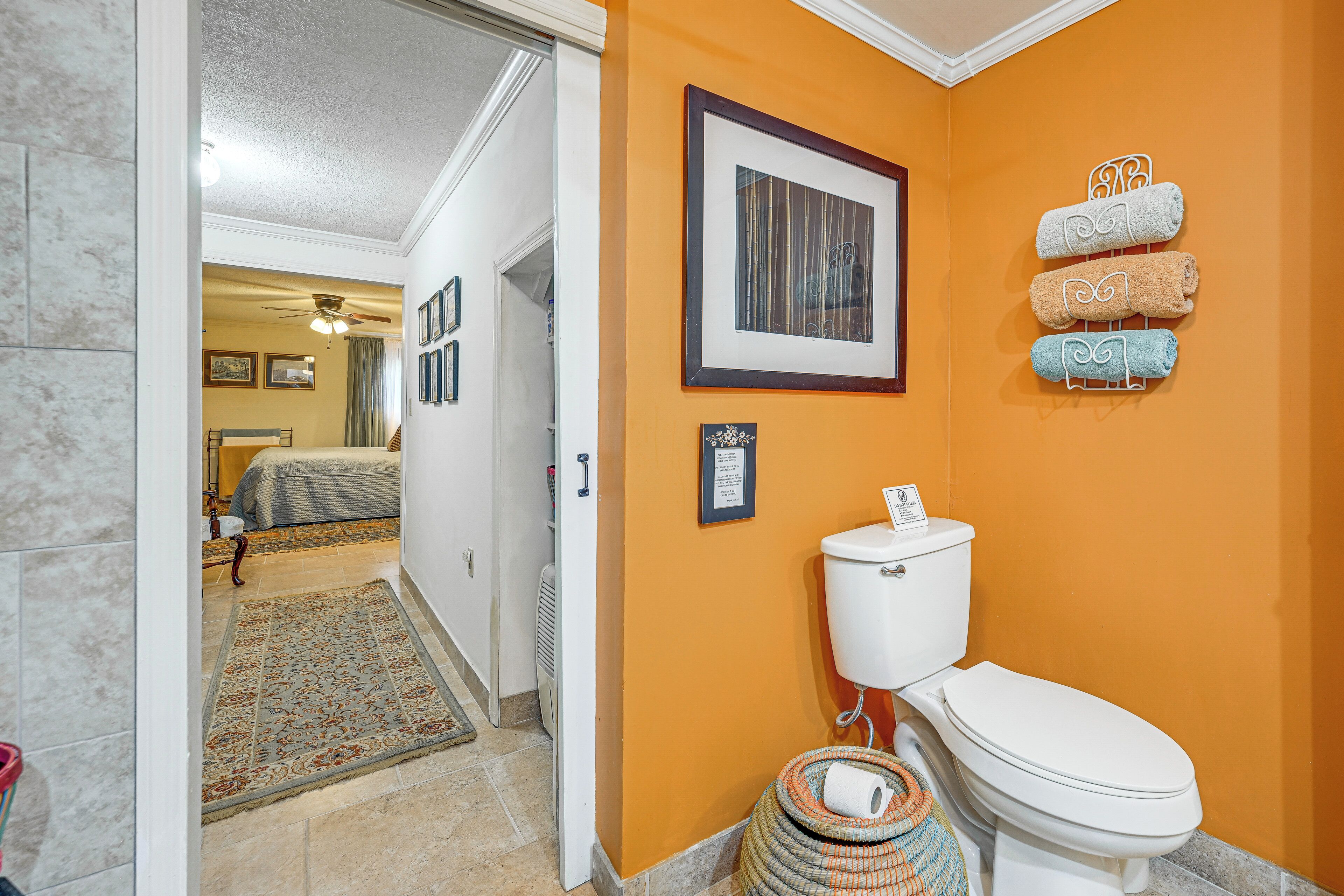 A bathroom with an orange wall, a white toilet, and a framed picture above the toilet. There are towels hanging on the wall and a basket with a roll of toilet paper.