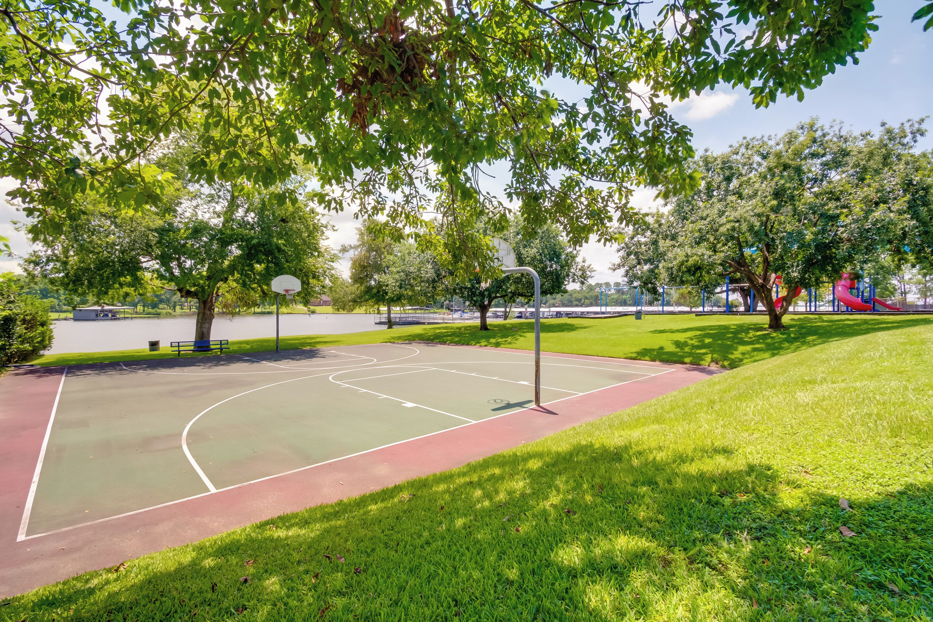 Una cancha de baloncesto al aire libre con superficie roja y verde, rodeada de césped y árboles.