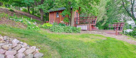 A cabin with a deck and a small porch, surrounded by trees and a grassy area.