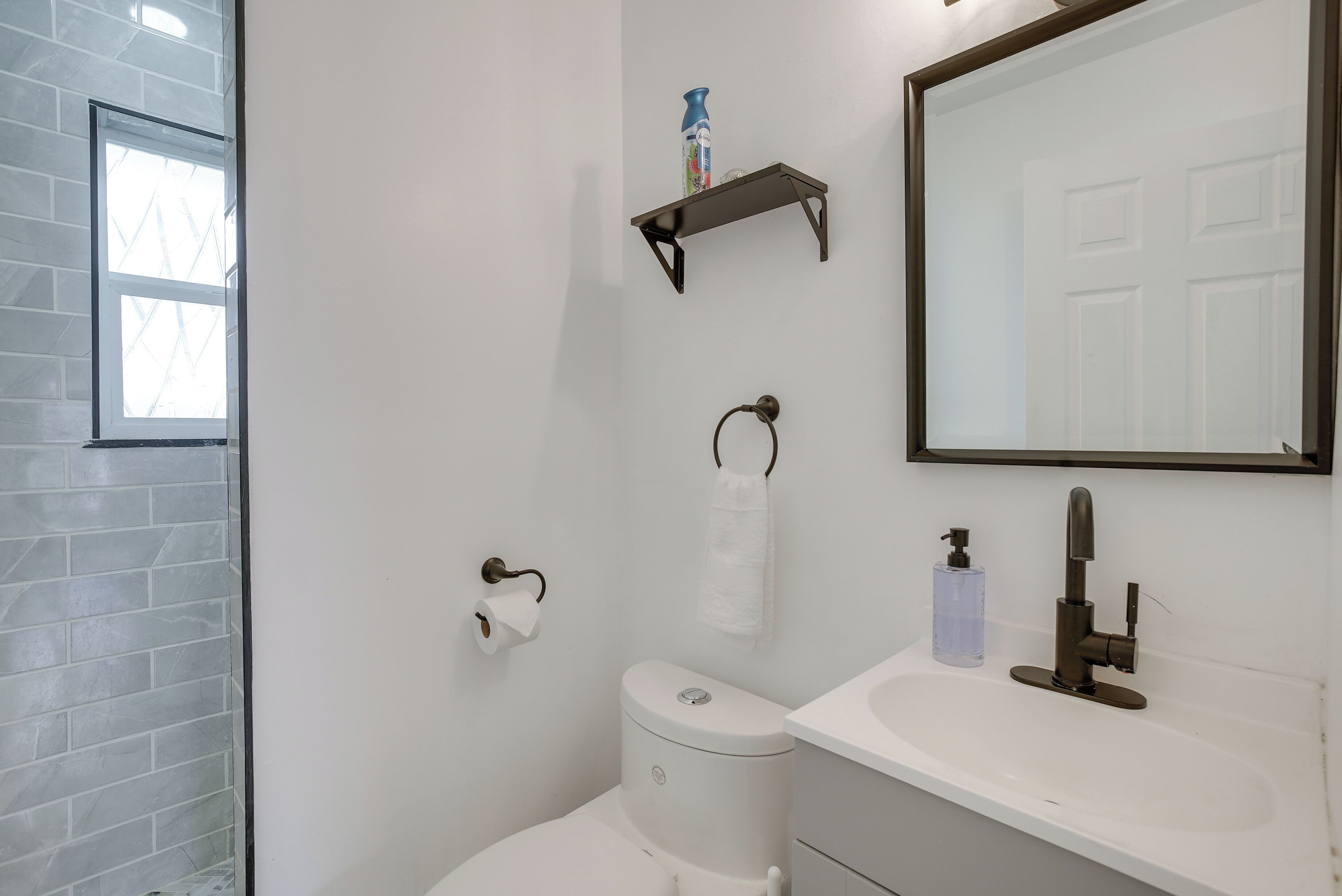 Un baño con inodoro blanco, lavamanos con espejo, toallero y ventana con vistas al exterior.