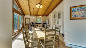 Een ruime eethoek met een houten tafel en stoelen, een keuken op de achtergrond en een groot raam met uitzicht op bomen.