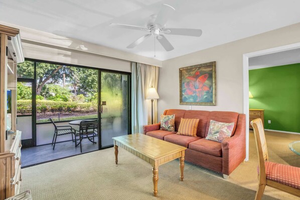 Un salon comprenant un canapé, une table basse, un ventilateur de plafond et une vue sur un espace extérieur accessible par une porte vitrée, où se trouvent une table et des chaises.
